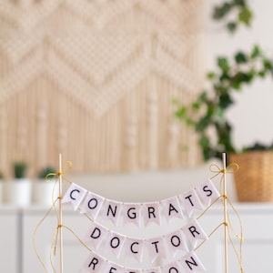 May include: White cake topper with a banner that reads "Congrats Doctor Reeson". The banner is made of white felt with black lettering and is strung on gold string.
