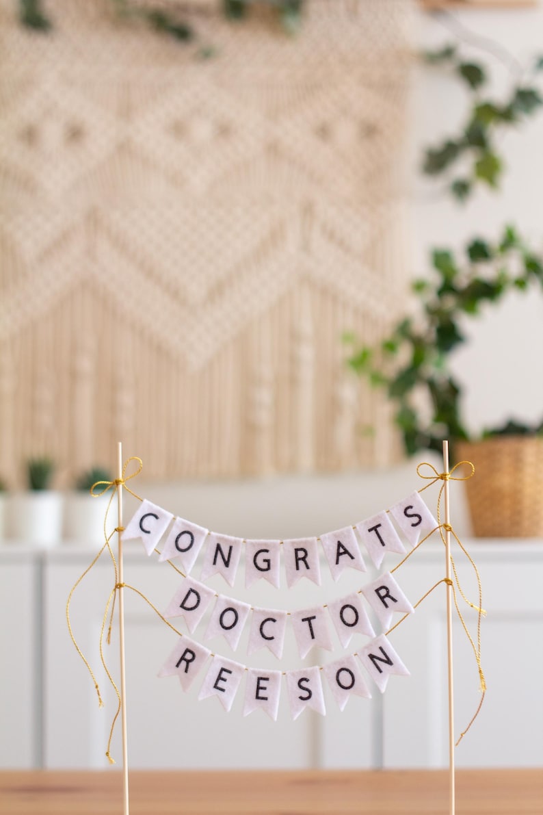 May include: White cake topper with a banner that reads "Congrats Doctor Reeson". The banner is made of white felt with black lettering and is strung on gold string.
