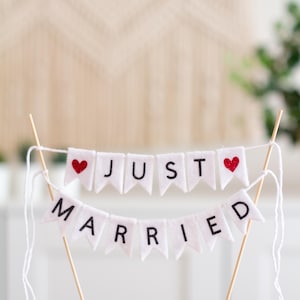 May include: A cake topper featuring two tiers of white felt pennant flags suspended between two wooden sticks. The top tier displays the word JUST with red glitter hearts on each end, and the bottom tier displays the word MARRIED in black letters.