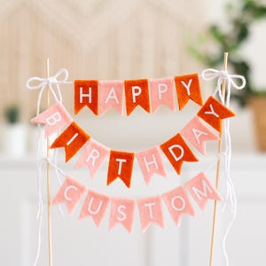May include: A cake topper featuring three tiers of felt pennant flags in alternating orange and light pink colors. The flags spell out "HAPPY BIRTHDAY CUSTOM" in white capital letters. The banner is suspended between two thin wooden sticks with white string.