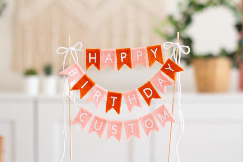 May include: A cake topper featuring three tiers of felt pennant flags in alternating orange and light pink colors. The flags spell out "HAPPY BIRTHDAY CUSTOM" in white capital letters. The banner is suspended between two thin wooden sticks with white string.