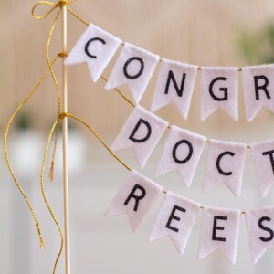 May include: White felt banner with gold string, spelling out "CONGRATS DOCS".