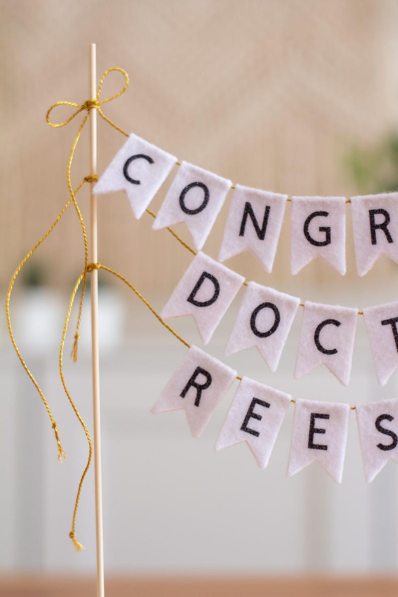 May include: White felt banner with gold string, spelling out "CONGRATS DOCS".