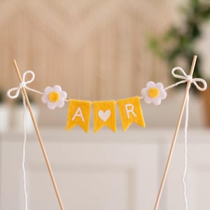 Può includere: Uno striscione di feltro giallo con le lettere "A" e "R" e un cuore bianco in mezzo. Lo striscione è infilato tra due bastoncini di legno con uno spago bianco e margherite di feltro bianco.