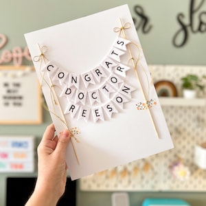 May include: A white card with a banner that reads "Congrats Doctor Reese" in black letters. The banner is made of small white flags with black letters and is strung together with gold string. The card is held in a person's hand.