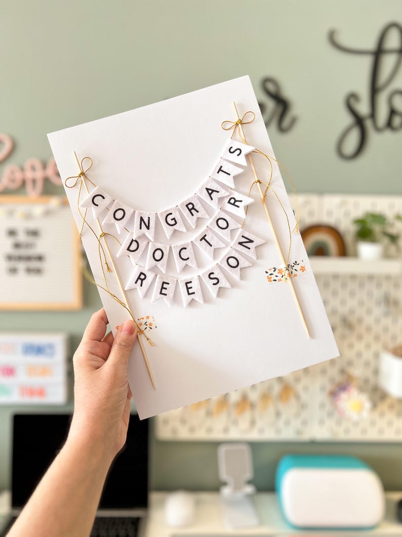 May include: A white card with a banner that reads "Congrats Doctor Reese" in black letters. The banner is made of small white flags with black letters and is strung together with gold string. The card is held in a person's hand.