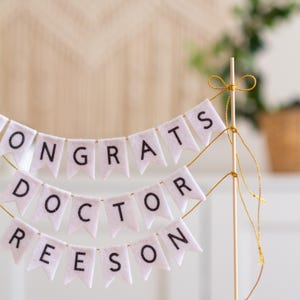 May include: White felt banner with the words "Congrats Doctor Reeson" strung on a wooden stick with gold string.