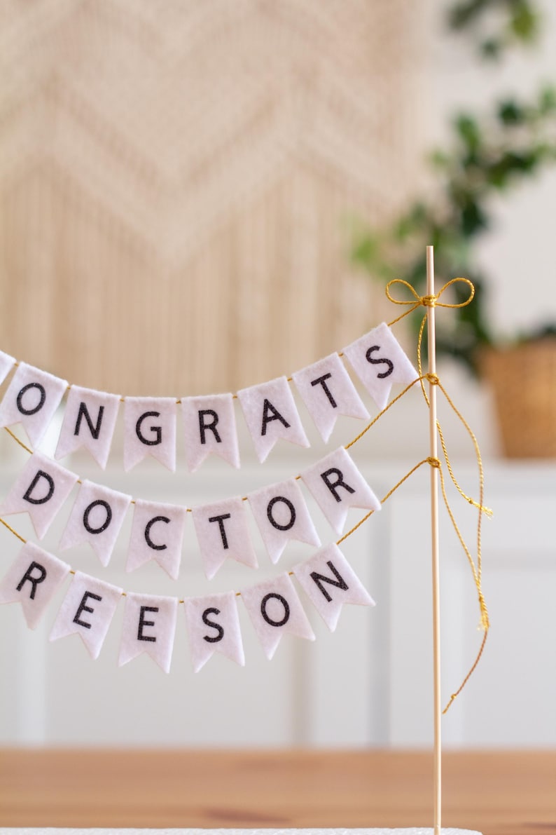 May include: White felt banner with the words "Congrats Doctor Reeson" strung on a wooden stick with gold string.