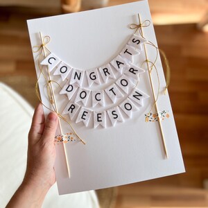 May include: White card with a felt banner that reads "Congrats Doctor Reeseon". The banner is strung with gold string and attached to two wooden sticks. The card is decorated with small floral patterned washi tape.