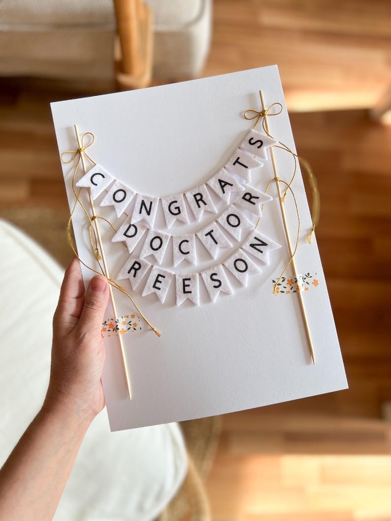 May include: White card with a felt banner that reads "Congrats Doctor Reeseon". The banner is strung with gold string and attached to two wooden sticks. The card is decorated with small floral patterned washi tape.