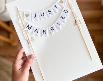May include: A white cake topper with the words "JUST MARRIED" in black letters on white pennant flags. The topper includes two wooden skewers and gold glitter heart accents. The topper is attached to a white card.