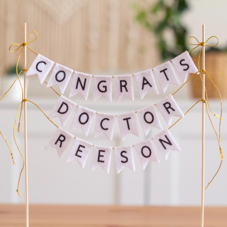 May include: White felt banner with gold string, reading "Congrats Doctor Reeseon" for a graduation cake topper.