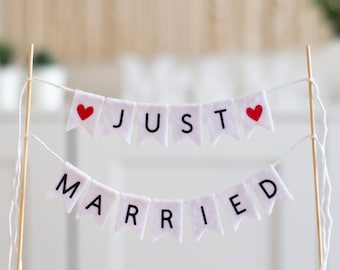 May include: A cake topper featuring two tiers of white felt pennant flags suspended between two wooden sticks. The top tier displays the word JUST with a red heart on each end. The bottom tier displays the word MARRIED in black letters.