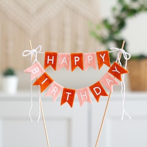 May include: A cake topper featuring two rows of felt pennant flags in alternating shades of burnt orange and light pink. The flags spell out HAPPY BIRTHDAY in white capital letters. The banner is suspended between two wooden sticks with white string.