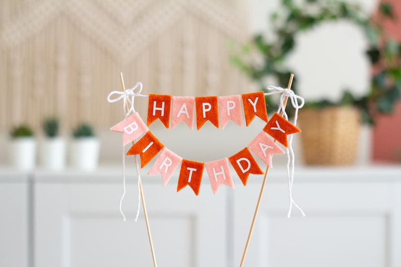 May include: A cake topper featuring two rows of felt pennant flags in alternating shades of burnt orange and light pink. The flags spell out HAPPY BIRTHDAY in white capital letters. The banner is suspended between two wooden sticks with white string.
