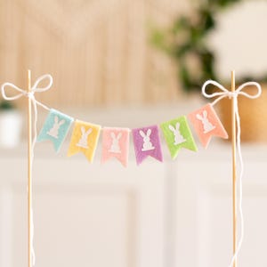 Op de afbeelding: Een pastelkleurig viltbanner met konijnenuitsnijdingen, perfect voor een lente- of paasfeest. De banner is aan een wit touwtje bevestigd en is bevestigd aan twee houten stokjes.