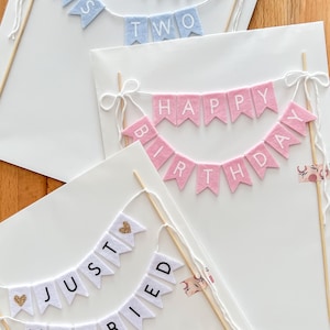 May include: Three cake toppers made of felt pennant flags attached to wooden skewers. One light blue banner reads GERARDO IS TWO. One pink banner reads HAPPY BIRTHDAY. One white banner reads JUST MARRIED with gold glitter heart accents.
