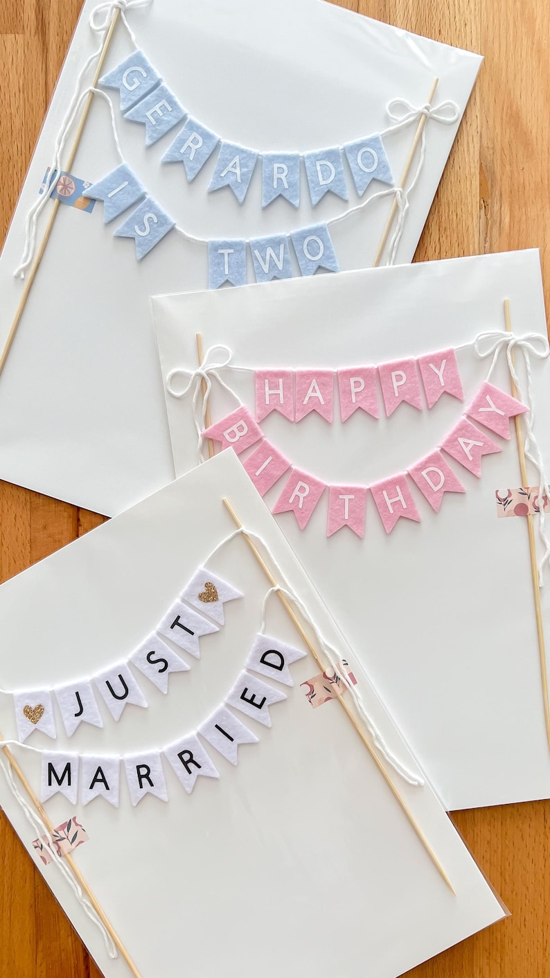 May include: Three cake toppers made of felt pennant flags attached to wooden skewers. One light blue banner reads GERARDO IS TWO. One pink banner reads HAPPY BIRTHDAY. One white banner reads JUST MARRIED with gold glitter heart accents.