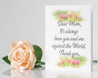 May include: A greeting card with the text "Dear Mom, It's always been you and me against the World. Thank you." is next to a peach-colored rose. The card has a watercolor floral design.