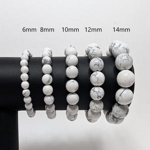 May include: Five white howlite bead bracelets of different sizes, displayed on a black stand. The bracelets are labelled 6mm, 8mm, 10mm, 12mm, and 14mm.