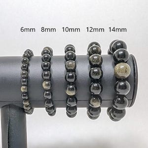 May include: Five black obsidian beaded bracelets with gold flecks, displayed on a black stand. The bracelets are sized 6mm, 8mm, 10mm, 12mm, and 14mm.