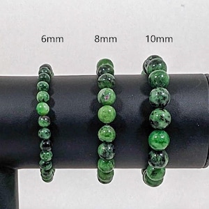 May include: Three green gemstone bracelets with different bead sizes: 6mm, 8mm, and 10mm. The bracelets are displayed on a black stand.