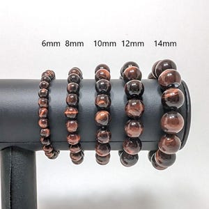 May include: Five red tiger's eye beaded bracelets of different sizes, 6mm, 8mm, 10mm, 12mm, and 14mm, displayed on a black stand.
