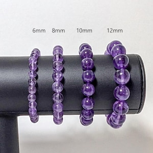 May include: Four amethyst gemstone bracelets displayed on a black stand. The bracelets are 6mm, 8mm, 10mm, and 12mm in size.