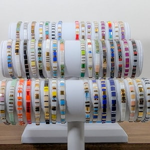 Peut inclure: Un présentoir de bracelets en perles colorées avec différents motifs et couleurs. Les bracelets sont disposés en rangées sur un support blanc.