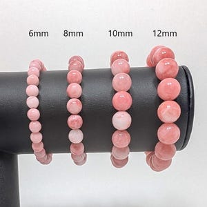 May include: Four pink gemstone bracelets displayed on a black stand. The bracelets are made with round beads in sizes 6mm, 8mm, 10mm, and 12mm.