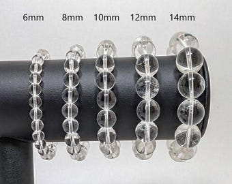 May include: Five clear quartz crystal bracelets of different sizes, displayed on a black stand. The bracelets are labeled 6mm, 8mm, 10mm, 12mm, and 14mm.