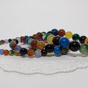 May include: Three colourful beaded bracelets made of agate stones. The bracelets are stacked on top of each other and feature a variety of colours, including blue, green, red, yellow, and black.