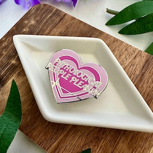Puede incluir: Un pin de esmalte en forma de corazón rosa y dorado con el texto "Pathological People Pleaser" en letras doradas.