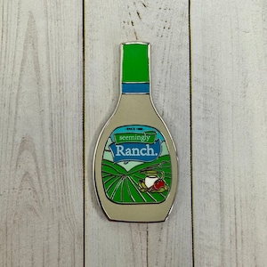 May include: A silver enamel pin shaped like a bottle of ranch dressing. The bottle is white with a green label that says "seemingly Ranch." The label also has a small green field with a red tomato, a white onion, and a white pitcher.