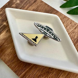 May include: A silver and black enamel pin featuring a UFO and a triangle with the text "Did you really beam me up?"