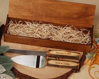 May include: A wooden box with a lid open, revealing a layer of shredded paper inside. The box contains a silver cake knife with two wooden handles. The handles are engraved with the text "January 11, 2019" and "Jacob & Jordan".