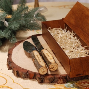 May include: A wooden box with a natural wood finish contains two silver cake knives with wooden handles. The handles are engraved with the words "Jenny & Justin" and a heart. The knives are sitting on a wooden slice.
