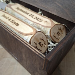 Può includere: Due fette di legno a forma di tronco con testo inciso e iniziali in una scatola di legno scuro. La fetta superiore recita "September 27, 2025" e la fetta inferiore recita "Jacob & Kayla".
