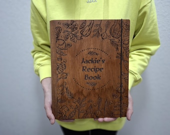 May include: A wooden recipe book with the text "Jackie's Recipe Book" in a circular design. The cover is decorated with engraved illustrations of vegetables and herbs. A black elastic band holds the book closed.