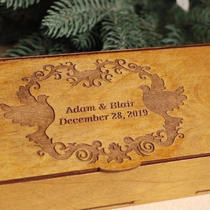 Op de afbeelding: Een houten doos met een gegraveerd ontwerp van twee duiven en een bloemige rand. De doos is gepersonaliseerd met de tekst "Adam & Blair December 28, 2019".