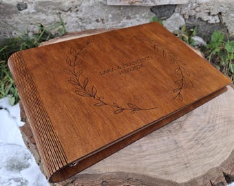 May include: A wooden photo album with a brown finish. The cover features a decorative engraved wreath with the text "Lara & Francesco 14.09.2024".