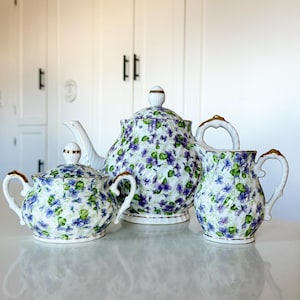 May include: A three-piece porcelain tea set featuring a teapot, sugar bowl, and creamer. The set is decorated with a floral pattern of purple and blue flowers on a white background, with gold trim.