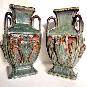 May include: Two ceramic vases with a green, brown, and red glaze. Each vase has a square base, a detailed relief design, two handles, and a flared top. The vases are displayed on a white surface.