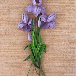 May include: A wall-mounted metal sculpture of three blooming purple iris flowers with yellow centers, two closed buds, and long green leaves. The floral arrangement features layered petals and stems, creating a dimensional botanical display.
