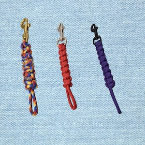 May include: Three paracord keychains with metal clasps. One is multi-colored, the second is red, and the third is purple. The keychains are displayed against a light blue denim background.