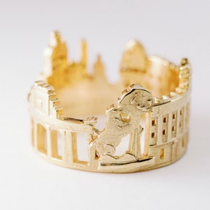 Könnte beinhalten: Ein goldfarbener Ring mit einem detaillierten Ausschnittdesign einer Stadtlandschaft. Der Ring zeigt eine prominente Löwenfigur und andere architektonische Elemente.