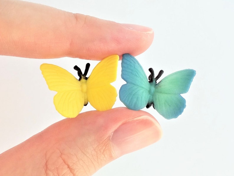 Peut inclure: Deux petits papillons en plastique, l'un jaune et l'autre bleu, avec des antennes noires. Les papillons sont tenus entre deux doigts.