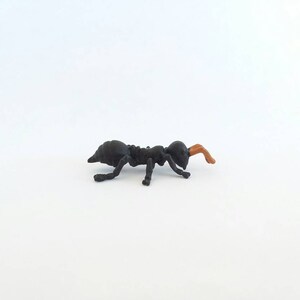 May include: A small, black ant figurine with a segmented body and legs. The ant's antennae are a light brown color. The figurine is set against a plain white background.