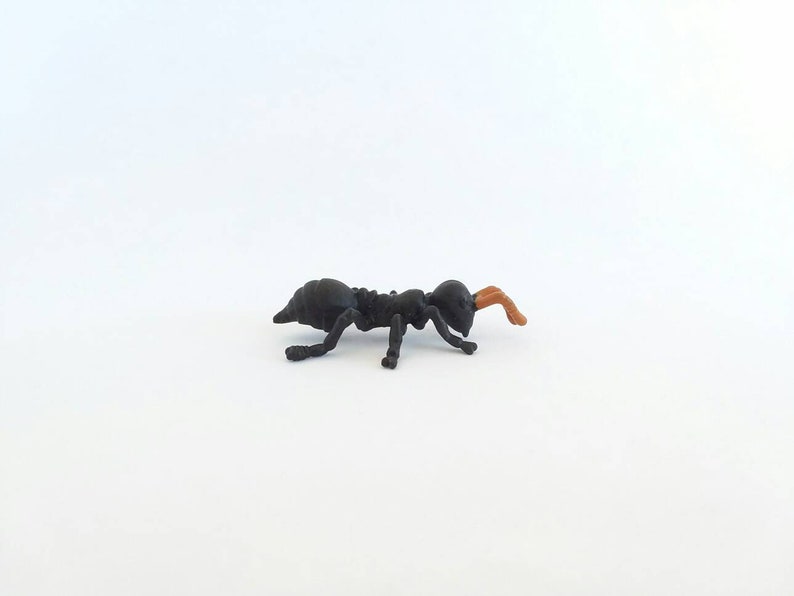 May include: A small, black ant figurine with a segmented body and legs. The ant's antennae are a light brown color. The figurine is set against a plain white background.