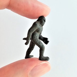 May include: A small, dark gray Bigfoot figurine, held between two fingers. The miniature figure has a detailed, textured surface and is posed in a walking stance. The background is plain white.
